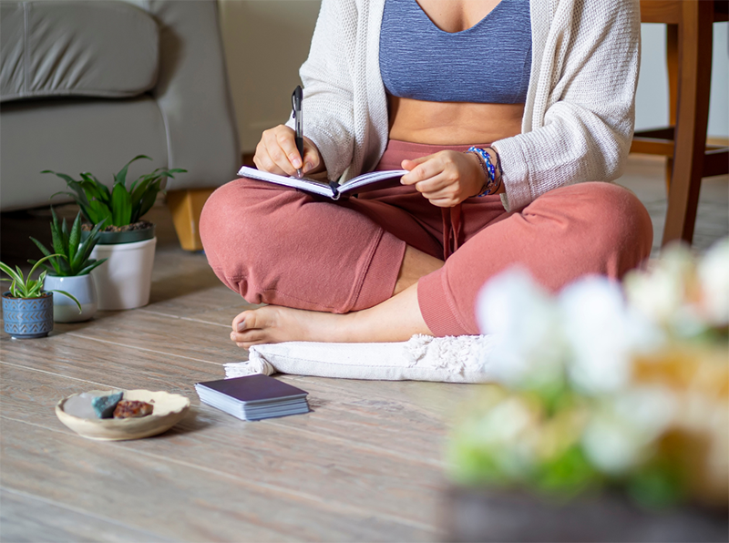 A person sitting on the floor next to plants. The person is practicing mindfulness techniques whilst writing in a journal.